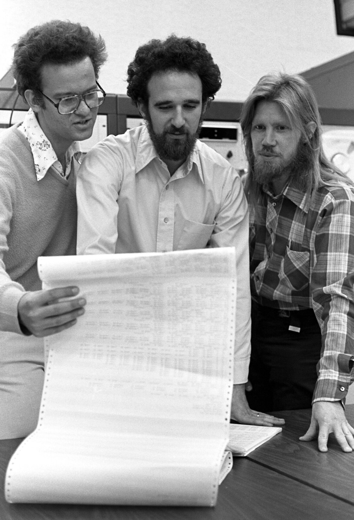 Photo en noir et blanc datant de 1977 montrant Merkle, Hellman et Diffie. Merkle tient un long et large papier listing édité par une imprimante matricielle. Les trois hommes lisent les informations écrites illisibles sur la photo.